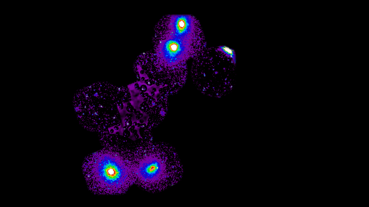 La imagen muestra un conjunto de manchas brillantes y coloridas sobre un fondo negro. Las manchas son principalmente moradas, con áreas de intenso brillo en el centro , que van del amarillo al verde y al azul. Estas manchas están conectadas por una tenue estructura morada, formando una forma irregular y extendida con manchas difusas en cada extremo